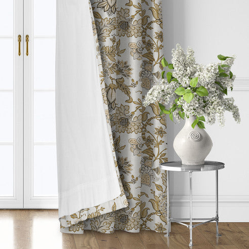 Bronte Gold floral drapery panel turned back to show its white lining beside a silver table with a vase of flowers.