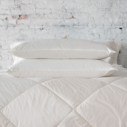 Three St. Genève Piatra Merino wool pillows stacked on a quilted bed against a white brick wall
