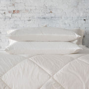 Three St. Genève Piatra Merino wool pillows stacked on a quilted bed against a white brick wall