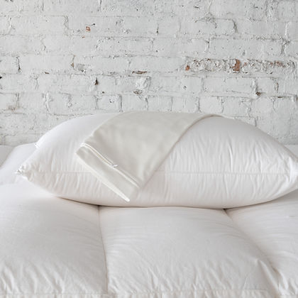 St. Genève Organic Pillow Protector covering fluffy white pillows against a white brick wall