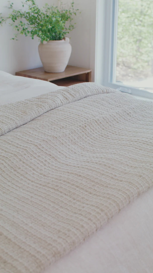 Spirit cream waffle cotton quilt folded back on a bed beside a window and a vase of greenery.