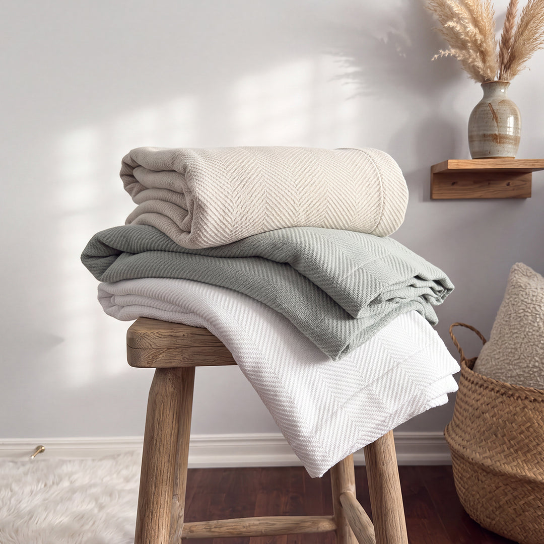 Cuddle Down Chevron cotton blankets folded and stacked on a wooden stool in white, sea glass, and pebble – 100% long-staple cotton
