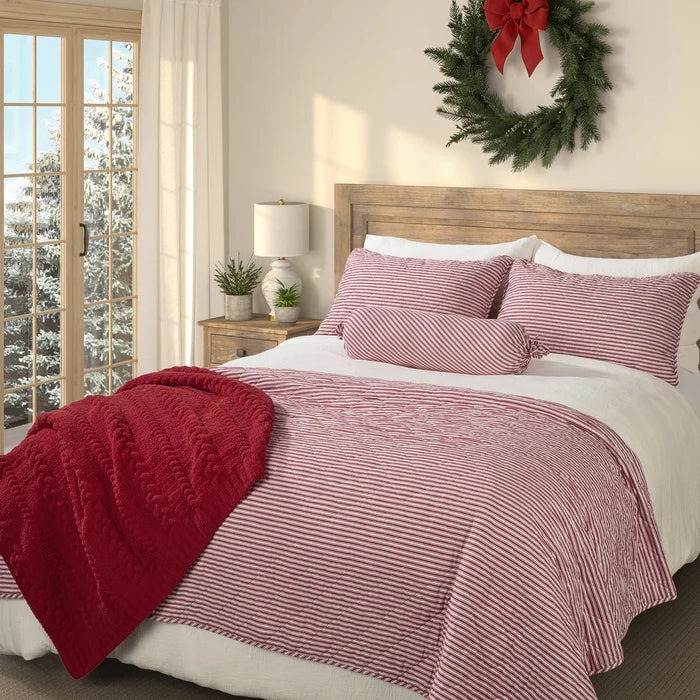 Holly red and white striped quilt on a bed with matching pillow shams, a red throw and a holiday wreath above the wooden headboard.