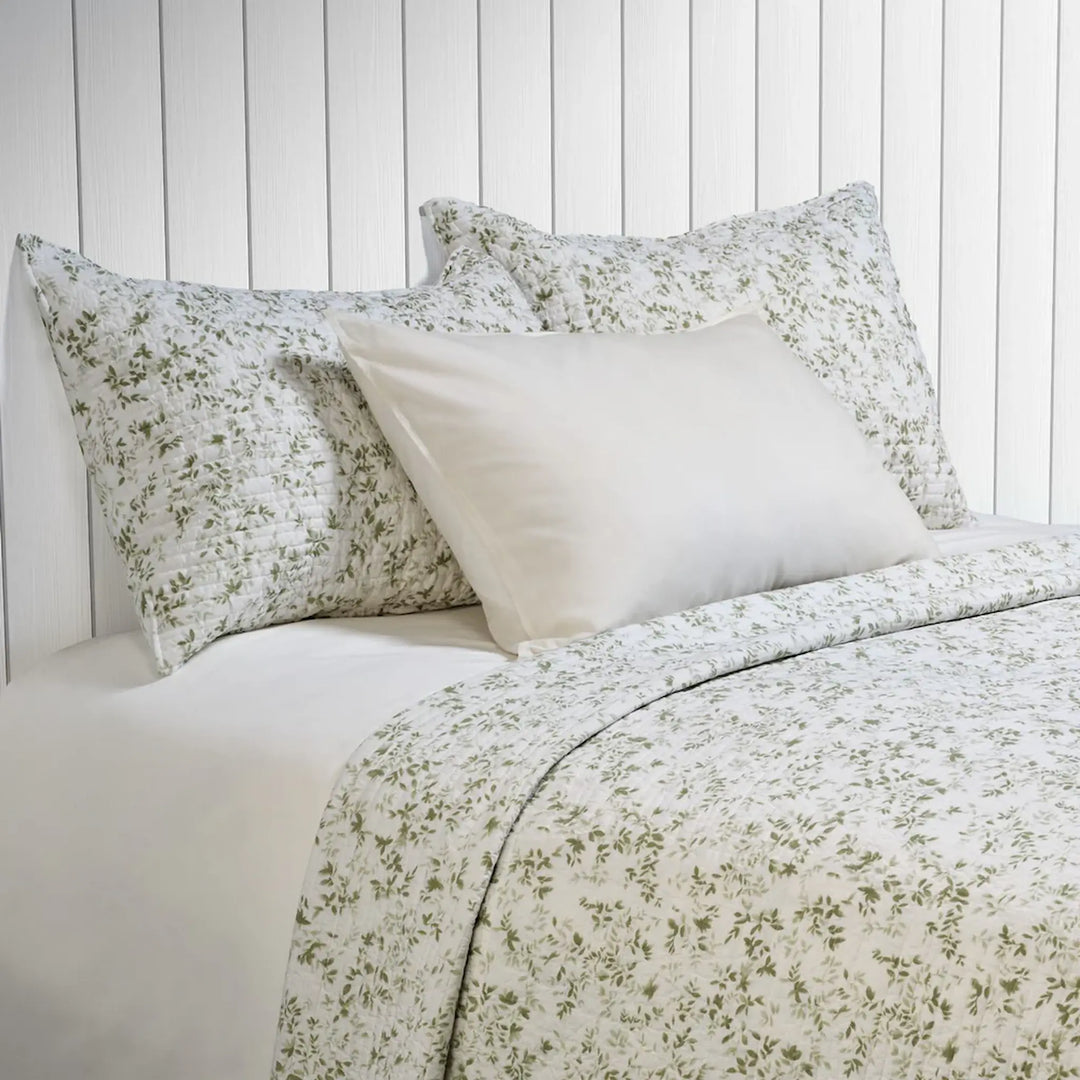 Marjolaine quilt set in white with sage green botanical foliage print, styled on a bed with matching pillow shams
