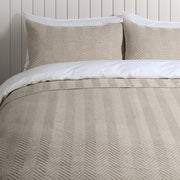 A beige textured quilted bedding set with matching pillow shams and white bedding, styled on a bed against a white wood-paneled wall.