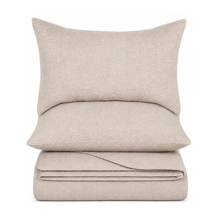 A stack of neutral beige home textiles, featuring a square throw pillow on top, a rectangular lumbar pillow behind it, and a folded sheet or blanket underneath, all shown against a plain white background.