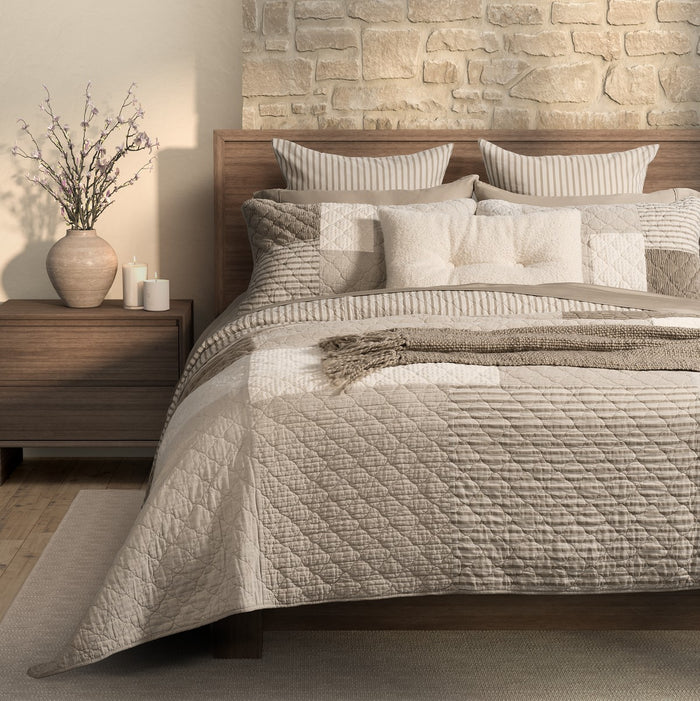 Brunelli Panorama natural and ivory patchwork quilt on a wooden bed with coordinating pillow shams and taupe striped accents.