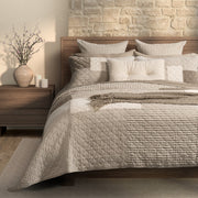Brunelli Panorama natural and ivory patchwork quilt on a wooden bed with coordinating pillow shams and taupe striped accents.